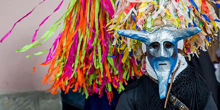 Una Maschera Cornuta dal volto azzurro e dal naso adunco accanto alla Fras, il ventaglio di nastri colorati