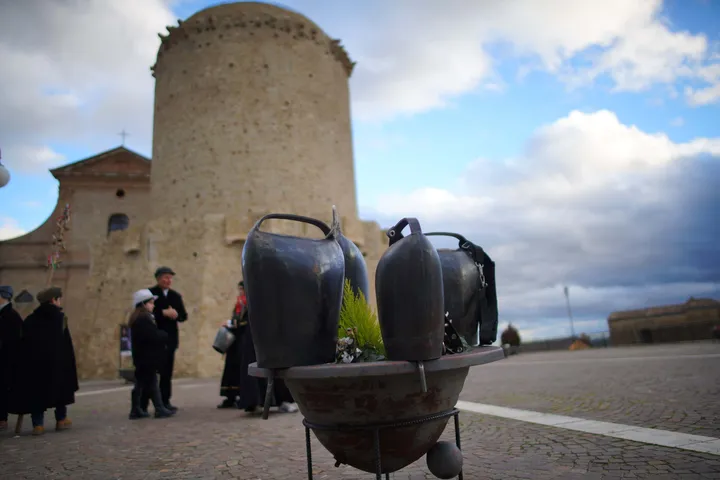 Due campanacci di bronzo appoggiati su una fontana; sullo sfondo la torre normanna di San Mauro Forte
